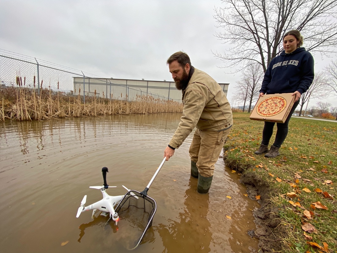 A man in waders reaching into a retention pond with a net to recover a drone while a woman holds a pizza box on the bank
